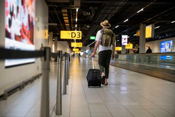 Low angle rear view of a young backpacker wearing a fedora, walking through the airport. He is ois off to catch his flight home.