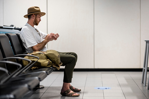 Side view of a young male backpacker sitting on a chair in the  airport, using his smart watch. He is getting ready to catch his flight home.