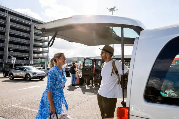 Mother and son packing up a rental van in France where they are vacationing to. They are standing in a car park at an airport in the sun.