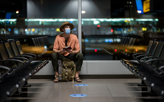 Side view of a young male backpacker sitting in an airport holding his passport and boarding pass. He is getting ready to catch his flight home and is wearing a protective face mask, to reduce to spread of COVID-19. It is dark outside the airport.