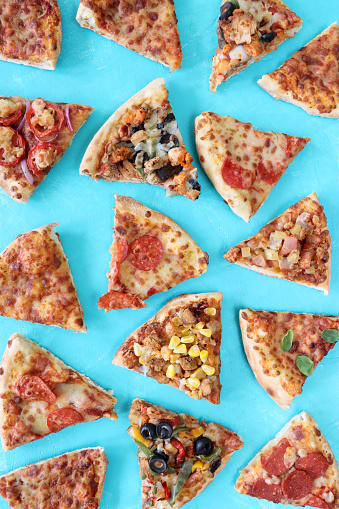 Stock photo showing elevated view of triangular slices of pizzas with different toppings including Hawaiian pizza (pineapple and ham), chicken and sweetcorn, pepperoni, vegetable (yellow and green bell pepper with black olives), feta, tomato and red onion, and Pizza Margherita (melted golden buffalo mozzarella cheese, rich tomato marinara sauce and fresh basil leaves).