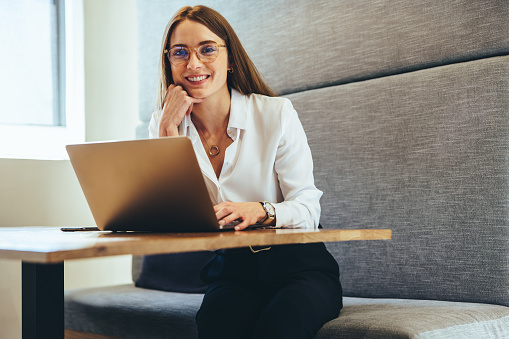 Professional businesswoman smiling at the camera while working in a modern workspace. Happy young female entrepreneur using a laptop while sitting alone on a couch.