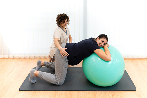 A pregnant caucasian woman doing pilates exercises with a ball with the help of her physiotherapist on the floor on a mat. Concept of physiotherapy pelvic floor treatment.