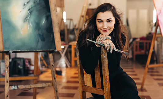 Self-employed young painter looking at the camera cheerfully while sitting on a chair in her workshop. Happy female artist holding a paintbrush with colour painted hands.
