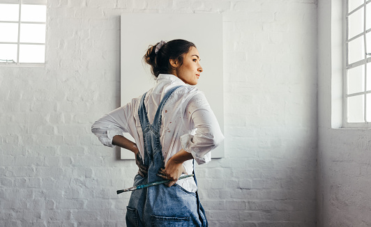 Painter looking away thoughtfully in her art studio. Rearview of a female artist contemplating new ideas while standing in front of a blank canvas. Creative young woman starting a new art project.