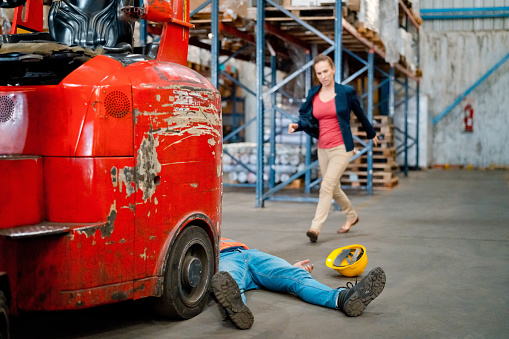 Man lying down on floor in front of in forklift in a warehouse. Female manager going to help him.