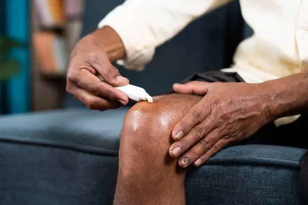 Close up of unrecognizable old man applying ointment cream for joint knee pain at home - concept of treatment or therapy for osteoarthritis and knee sprain.