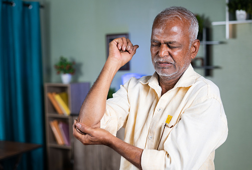 Indian senior old man exercising by strenching hand due to elbow joint pain at home - concept of suffering joint pain and sprain injury