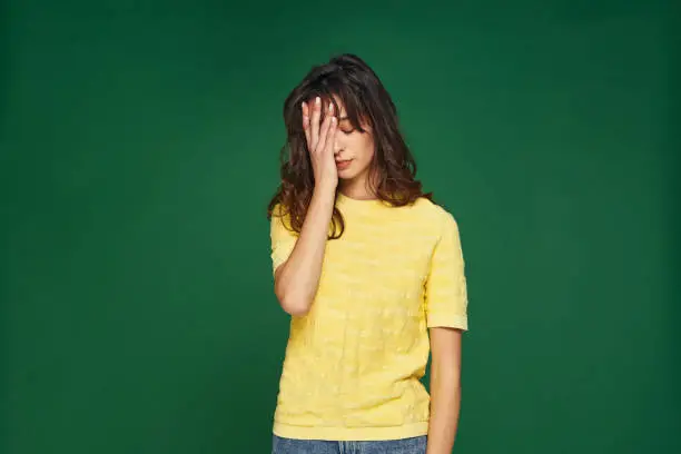 Photo of Facepalm gesture. Stressed tired young girl cover face by hand feel sorrow, shame for mistake on green studio background