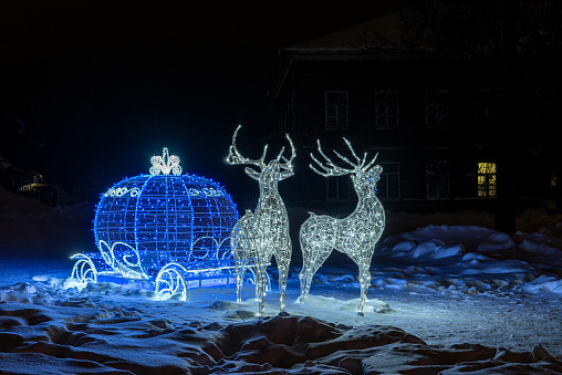 Glowing Reindeer harnessed to a carriage in the form of a Christmas ball. Street festive installation decorated with garlands.