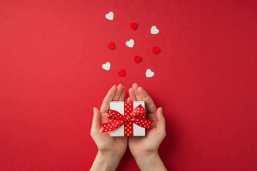 First person top view photo of valentine's day decorations female hands holding small white giftbox with red dotted ribbon bow on palms and hearts on isolated red background with copyspace