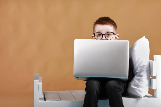 istock schoolboy in glasses using laptop sitting on bed. Child doing school homework and seriously and tired looking on camera. Online learning, distance lesson, education with online tutor at home isolation 1364227207