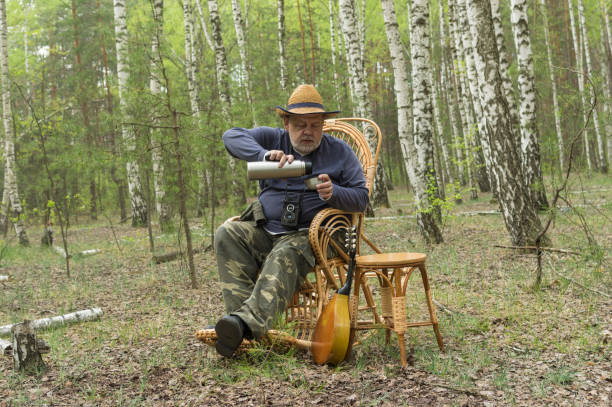 Bearded Caucasian  senior camper is having rest in birch forest, sitting on a wicker rocking chair and drinking tea from thermos bottle