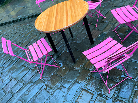Stock photo showing close-up image of circular wooden table with black, metal legs surrounded by brightly coloured, pink painted, folding metal chairs on wet cobbled road, as part of bistro cafe outdoor seating arrangements.