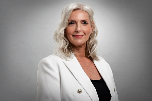 Studio portrait of grey haired senior woman looking at camera and smiling while standing at isolated grey background. Copy space.