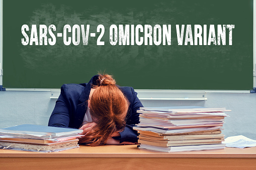 Teacher on a green blackboard background with the text new covid variant Omicron. Woman school teacher crying at her desk with books