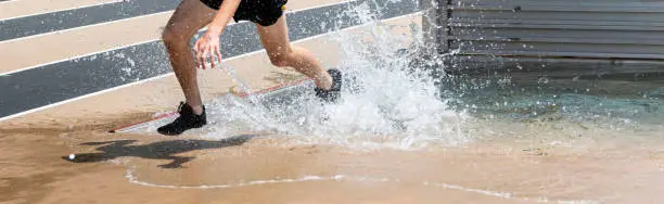 A high school boy is splashing in the water as he is exiting the steeplechase water pit during a track race.