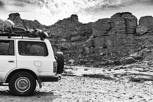 Left half rear side of a four wheels drive car parked in the reg with dry stones in the Sahara in black and white contrasted stone mountain with cloudy sky in the background.