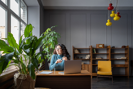Happy freelancer woman relaxing from work in coworking space or library dreamy looking in window after successful video conference meeting with client, employer or team leader. Joyful writer female