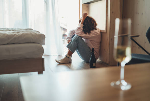 mulher triste sentada no chão do quarto com uma garrafa de álcool. taça de vinho desfocada na mesa de primeiro plano. problemas de saúde mental e alcoolismo conceito imagem. - abuso de álcool - fotografias e filmes do acervo