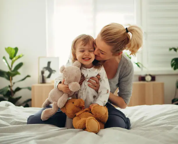 Beautiful blonde woman is playing with her cute baby girl on the bed. She is sitting behind her, hugging and kissing her. The girl is holding stuffed animal toys. Both of them are smiling. They are still in their pyjamas.