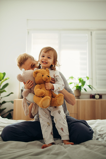 Beautiful blonde woman is playing with her cute baby girl on the bed. She is sitting behind her and hugging her. The girl is standing on the bed holding stuffed animal toys. Both of them are smiling. They are still in their pyjamas.