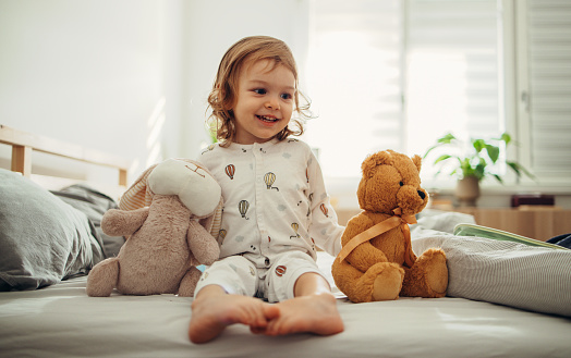 Portrait of the baby girl sitting barefoot in her pyjamas on the bed in the morning. She is playing with stuffed animals and smiling.