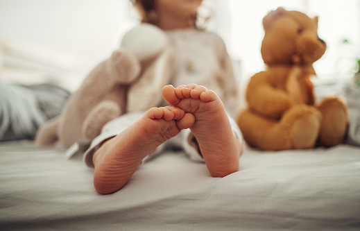 Anonymous child is sitting on the bed barefoot. Their feet are in focus. There are stuffed animals  visible in the back.