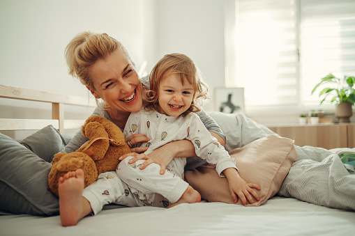 Portrait of the baby girl sitting barefoot in her pyjamas on the bed in the morning. She is sitting with a teddy bear and smiling while her mom is cuddling with her. Both of them are smiling.
