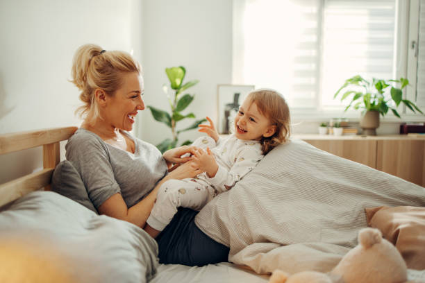 feliz madre e hija acurrucadas en la cama por la mañana - playing playful baby contemporary fotografías e imágenes de stock