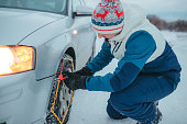 Young male adult putting on snow chains on his car tires during a snowfall.