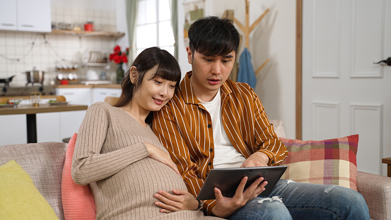 happy asian pregnant couple shopping online together for baby stuff with an electronic pad on the sofa in the living room at home