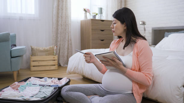 istock expectant woman is writing a memo in her notebook while thinking and preparing clothing for her upcoming newborn at bright home interior. 1364323622