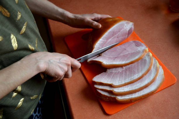 jovem corta presunto magro em fatias grossas com uma faca. placa de corte de plástico laranja. o presunto tem uma cor rosa bonita e a borda ousada com a pele caracteriza a carne de porco praga, verde - knife edge - fotografias e filmes do acervo