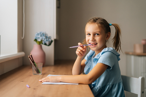 Cheerful pensive primary little child girl doing homework and holding pen against mouth sitting at home table by window, looking at camera. Thinking dreaming preschool kid studying at bedroom.