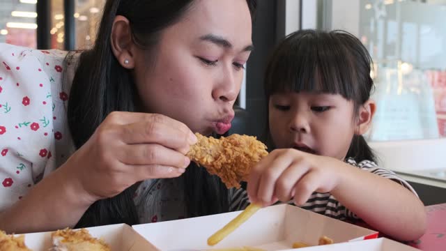 Cute little Asian girl eating fried chicken and french fries together with a beautiful mother at a fast food restaurant.
