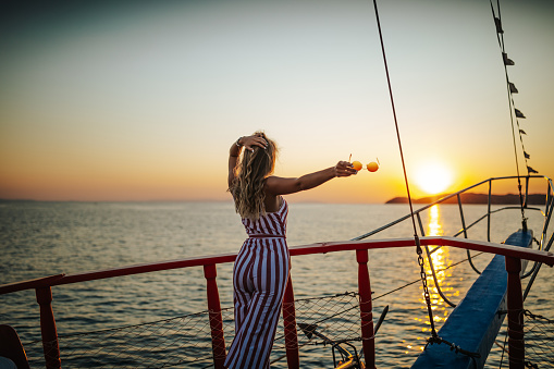 Beautiful young woman standing on boat prow and admiring sunset above the sea