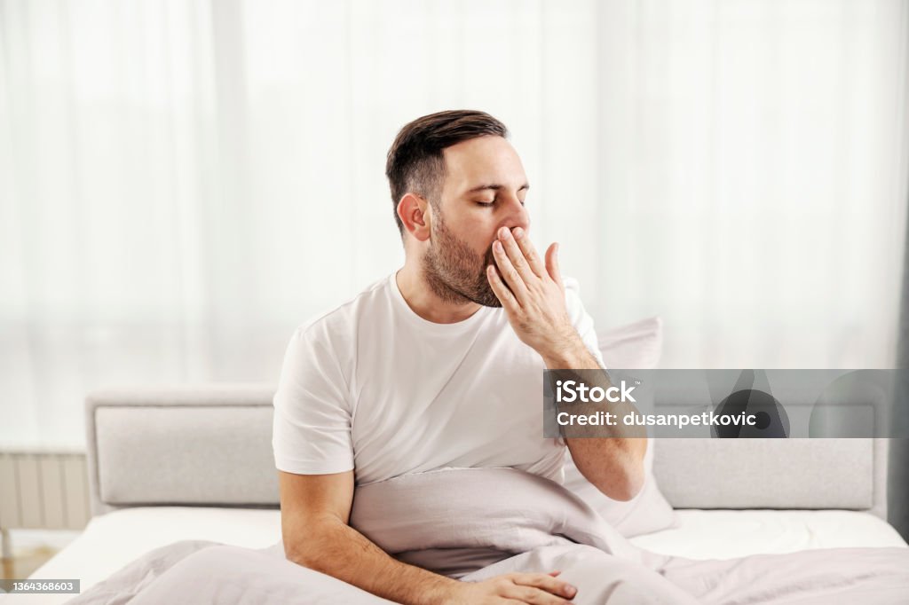 A morning man sitting in the bed, yawning and waking up at his cozy home. Yawning Stock Photo