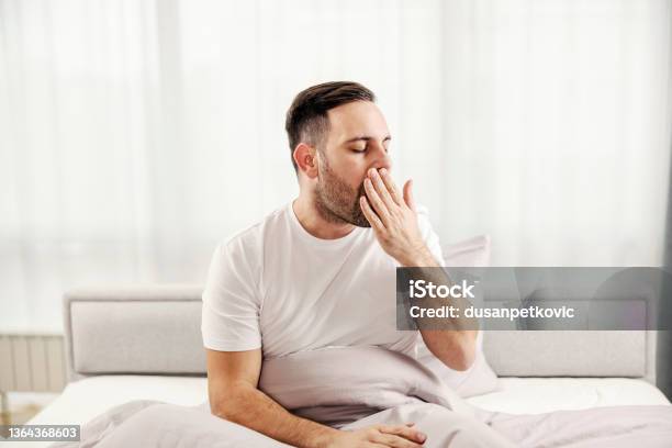 A Morning Man Sitting In The Bed Yawning And Waking Up At His Cozy Home Stock Photo - Download Image Now