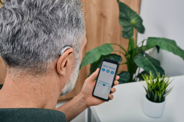 Hearing impaired mature man adjusts settings for his BTE hearing aid via smartphone. Hearing aids, deafness treatment, innovative technologies at audiology stock photo