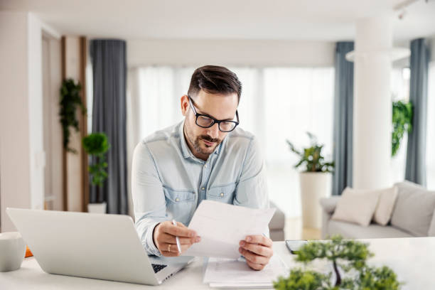 istock A serious man looking at paperwork and bills while standing next to a laptop at his home office. 1364368704