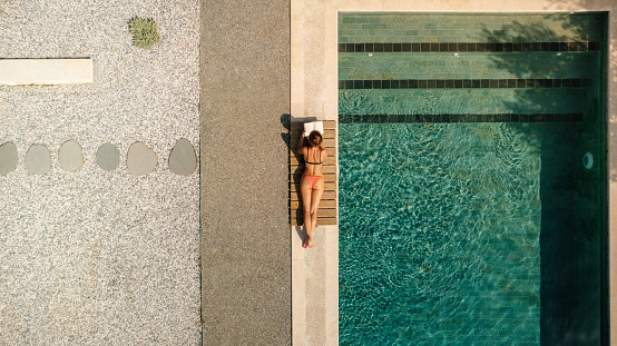 Photo of a young woman enjoying the sunny day by the pool and reading a book
