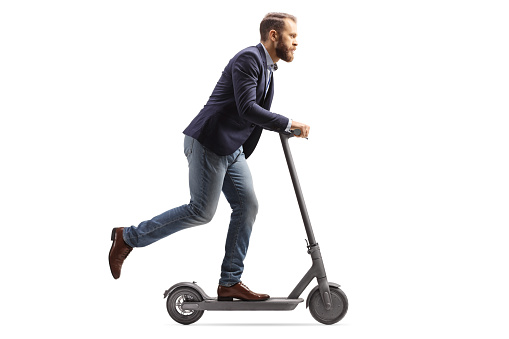 Full length profile shot of a young man in suit and jeans riding an electric scooter isolated on white background
