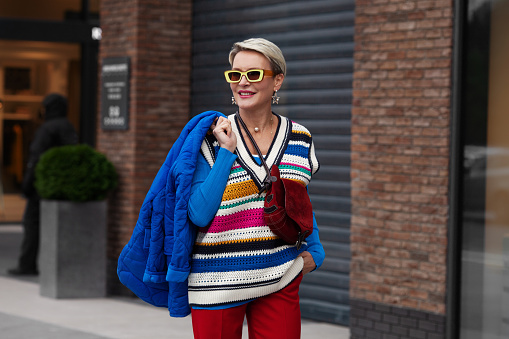 Smiling Happy Model lleva chaqueta y suéter azules, chaleco a rayas, pantalones rojos y gafas de sol amarillas. Moda Street style otoño o primavera. Mujer posando afuera de la tienda de ropa photo