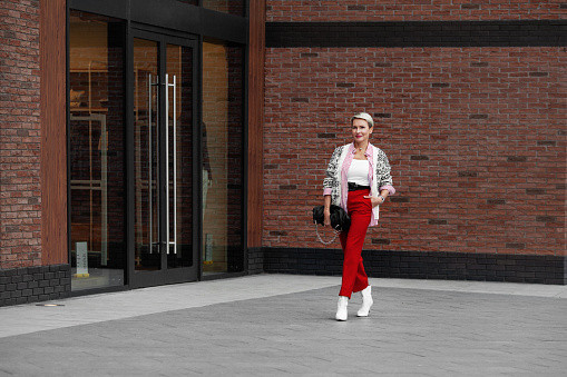 Autumn or Spring street style trend Clothes. Bright outfit for business woman walking confidently down the street. Model wearing sweater, shirt, red pants, white leather boots. Copy space