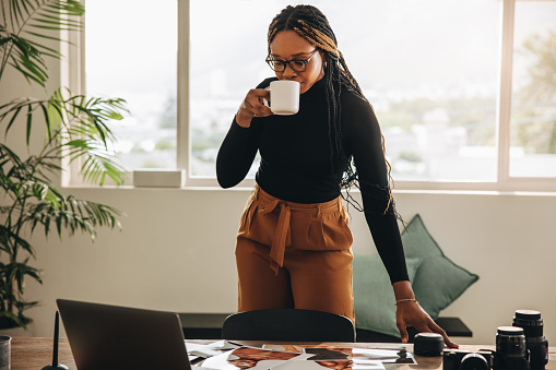 Self-employed young woman drinking coffee in her home office. Creative young woman standing at a desk with photographic equipment. Female photographer taking a coffee break while working on a new project.
