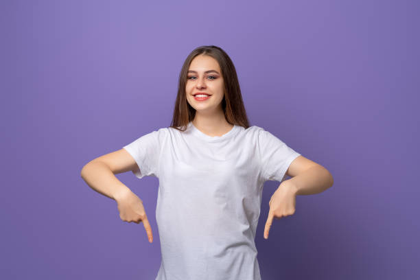 istock Smiling beautiful woman points at bottom, has happy facial expression, wears blank white t shirt, isolated over purple background. Look downwards, your promo 1364376227