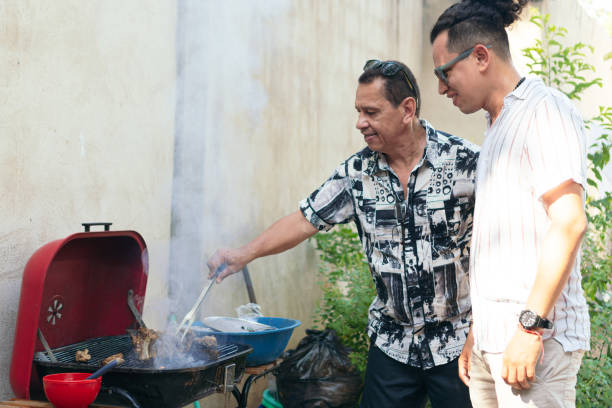 uomo anziano che prepara carne su un barbecue per il pranzo della sua famiglia nel cortile di casa. suo figlio è in piedi accanto a lui. - automovil foto e immagini stock