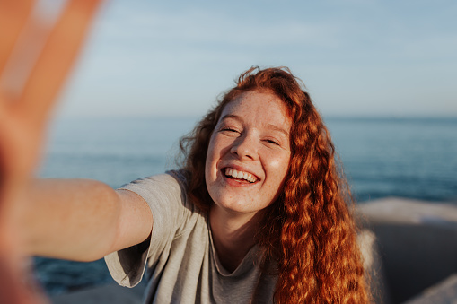 Cheerful young woman taking a selfie while standing next to the sea. Carefree young woman smiling happily at the camera. Outdoorsy young woman having fun in the summer sun.