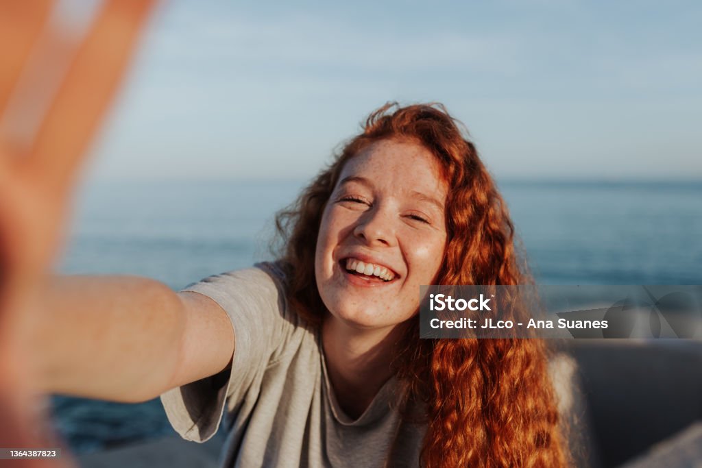 Cheerful young woman taking a selfie next to the sea Cheerful young woman taking a selfie while standing next to the sea. Carefree young woman smiling happily at the camera. Outdoorsy young woman having fun in the summer sun. Portrait Stock Photo